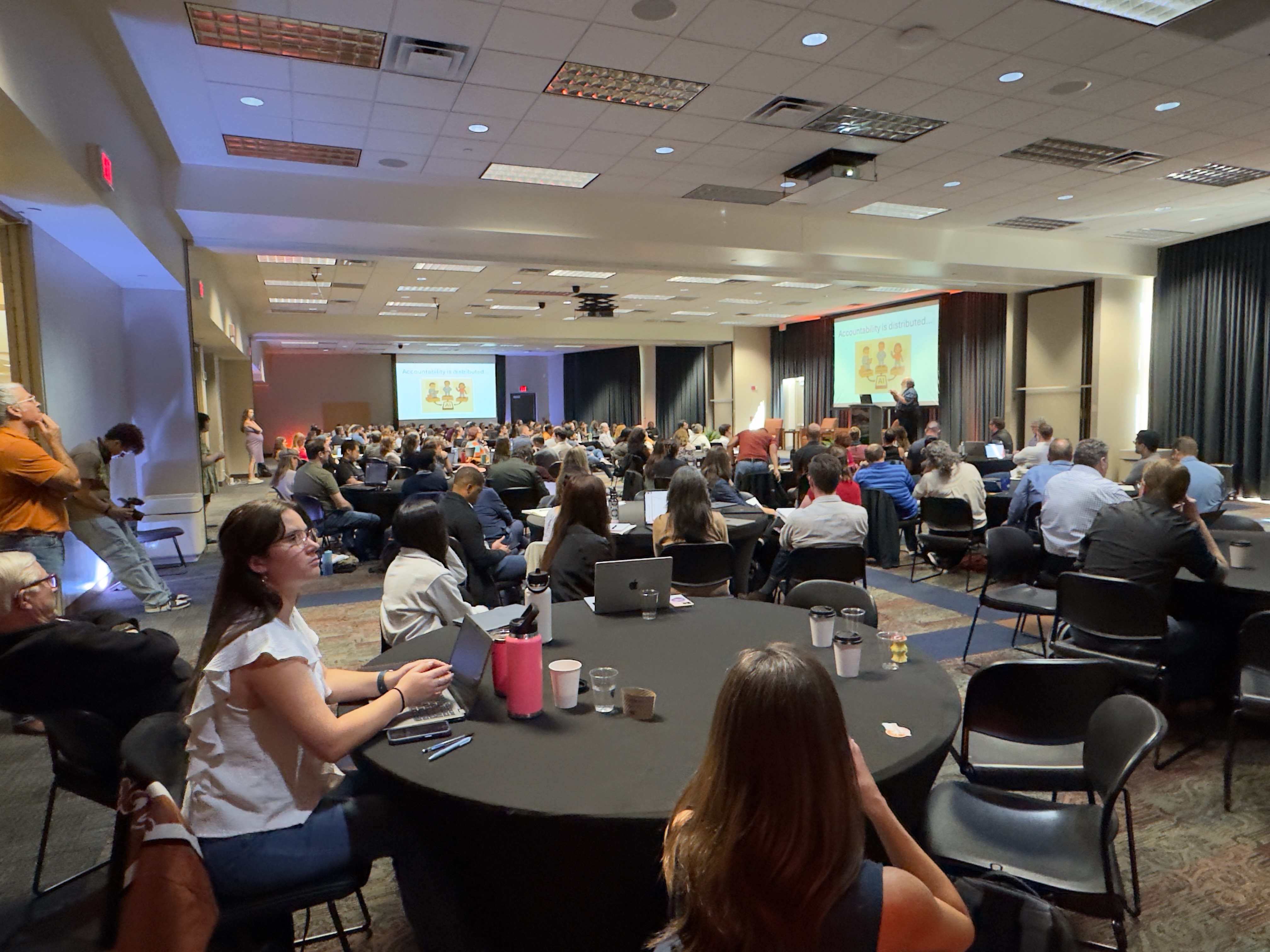 Audience members gathered at round tables during AI in a Day while a speaker presents on stage.